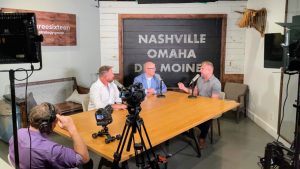 Three middle aged white men speaking into podcast microphones while sitting in a podcast studio in Omaha