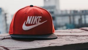 a red & black baseball hat with a white nike logo sits on the cement. There is a city skyline in the background.