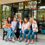 five women wearing jeans and sweaters sitting on a black bench outside of a floral and gift shop in Omaha