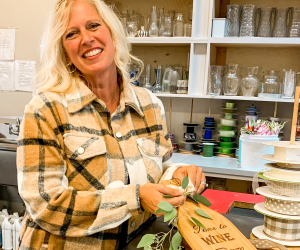 a white women with blonde hair with a yellow flannel shirt is holding a wooden carving board at an omaha gift shop