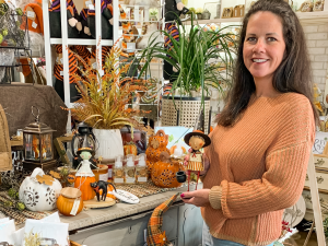 a white women with dark hair is wearing an orange sweater and holding a small scarecrow figurine at a gift shop in omaha.