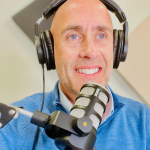 a handsome middle aged white male with a blue sweater wearing podcast headphones and sitting in a podcast booth.