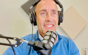 a handsome middle aged white male with a blue sweater wearing podcast headphones and sitting in a podcast booth.