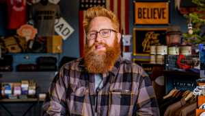 A white man with a red beard and glasses is wearing a flannel shirt. He is standing in a men's store in omaha, nebraska.