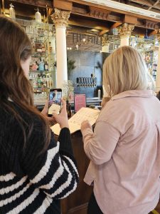 two women next to each other. One women has brown hair and a striped sweater white and blue, she is capturing a video of the other women looking through the menu. The other women is blonde and wearing a light pink flannel