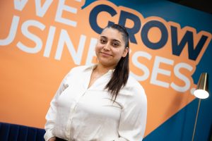 a woman in a white blouse and dark hair standing in front of an orange wall that says we grow businesses. 