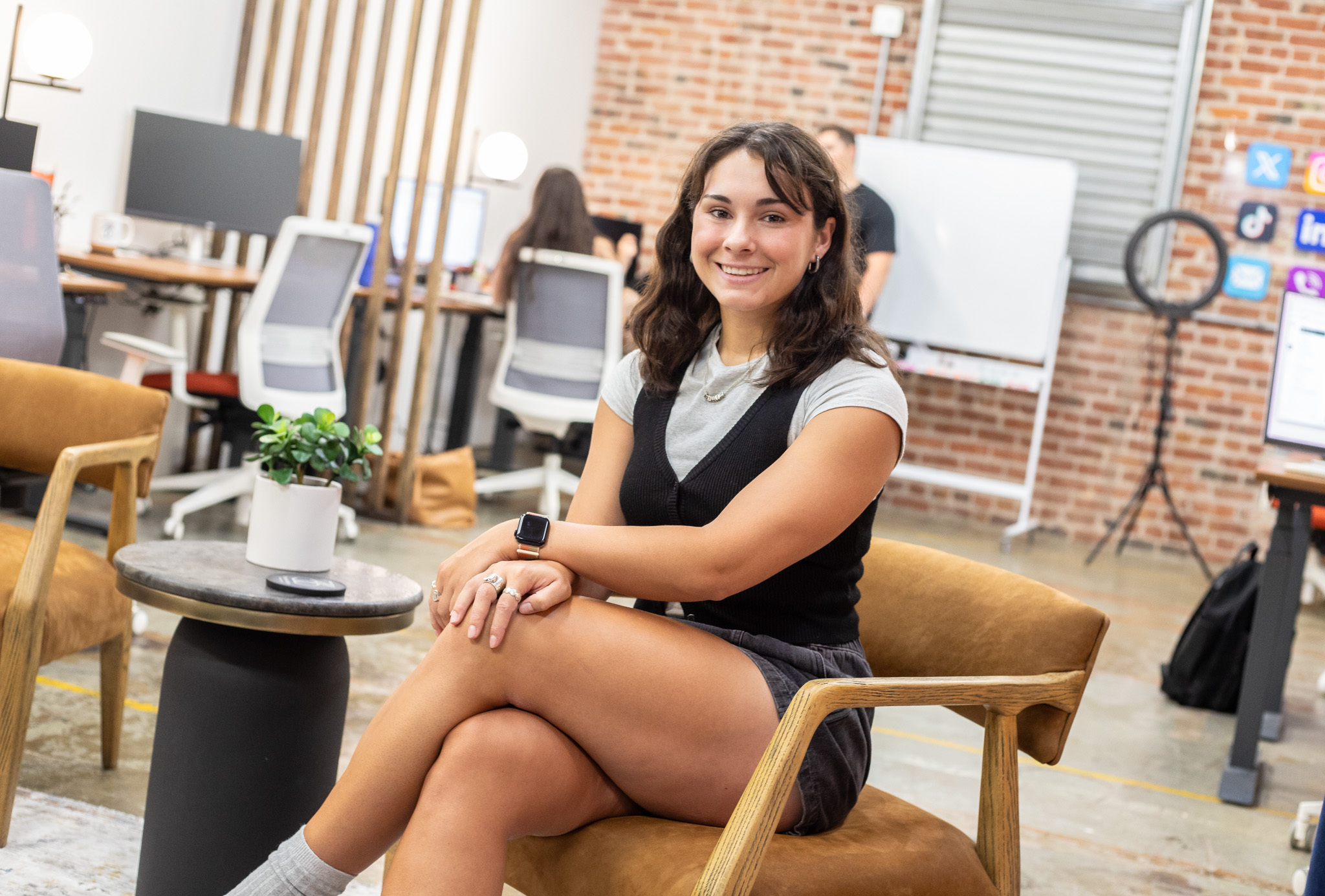 A woman in black shorts, white shirt, and black vest sits on a leather chair in a trendy, urban, industrial content house. 